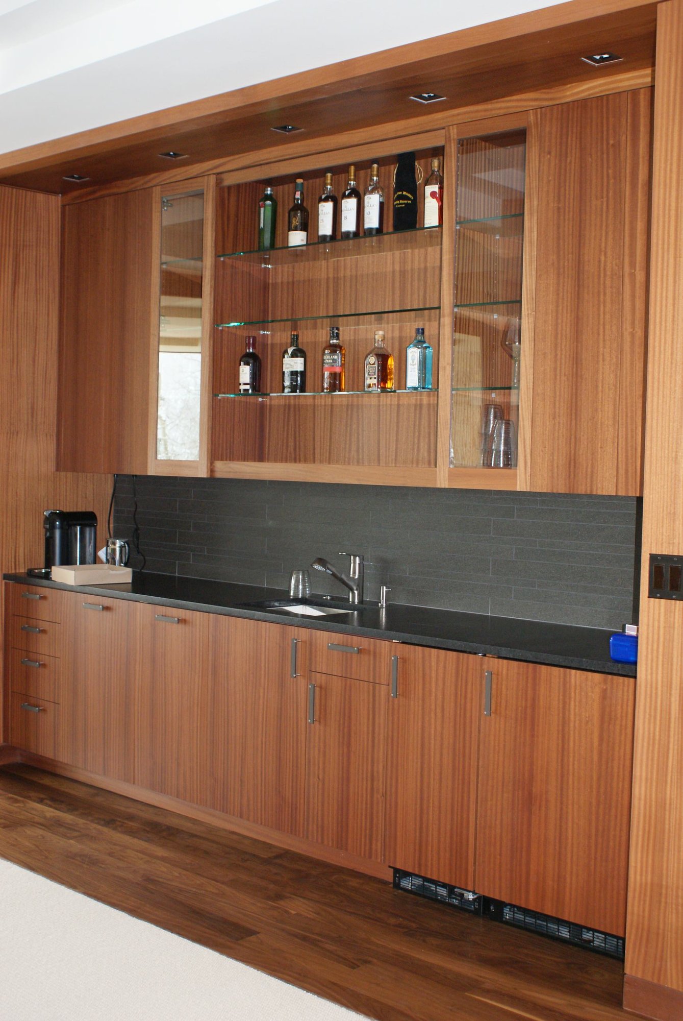 Mid-sized contemporary single-wall dark wood floor wet bar with an undermount sink, flat-panel cabinets, medium tone wood cabinets, black backsplash and solid surface countertops, by Jensen Design in Telluride, CO