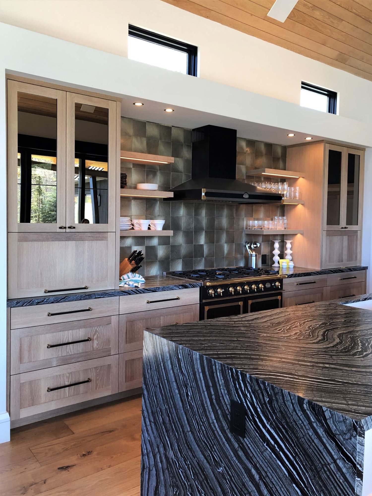 Mid-sized trendy galley medium tone wood floor, brown floor and wood ceiling open concept kitchen with a single-bowl sink, shaker cabinets, light wood cabinets, metallic backsplash, black appliances, an island and black countertops, by Jensen Design in Telluride, CO
