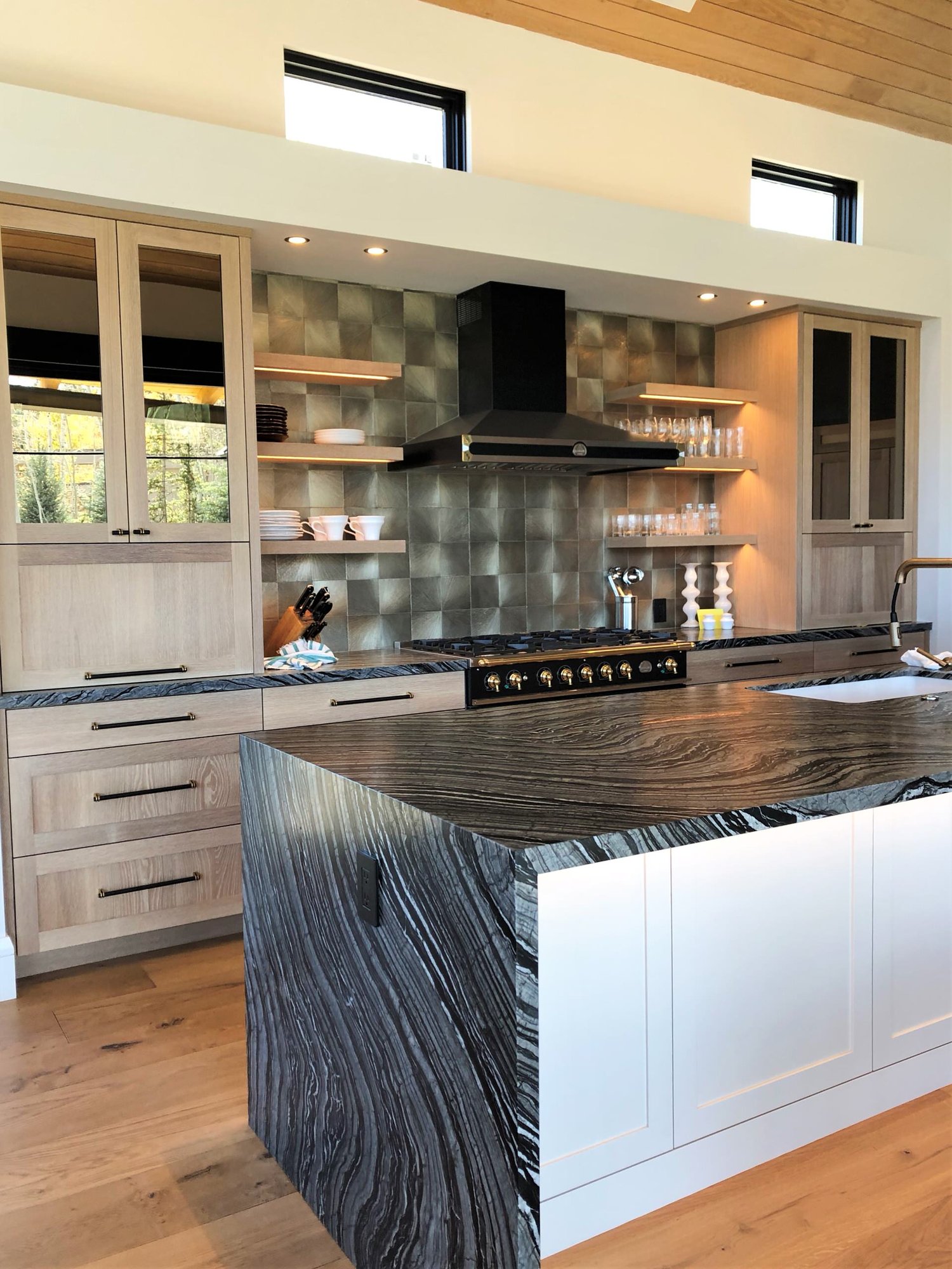 Mid-sized trendy galley medium tone wood floor, brown floor and wood ceiling open concept kitchen with a single-bowl sink, shaker cabinets, light wood cabinets, metallic backsplash, black appliances, an island and black countertops, by Jensen Design in Telluride, CO