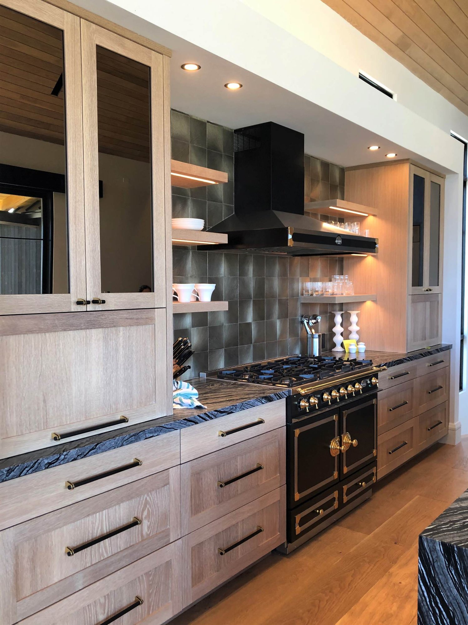Mid-sized trendy galley medium tone wood floor, brown floor and wood ceiling open concept kitchen with a single-bowl sink, shaker cabinets, light wood cabinets, metallic backsplash, black appliances, an island and black countertops, by Jensen Design in Telluride, CO