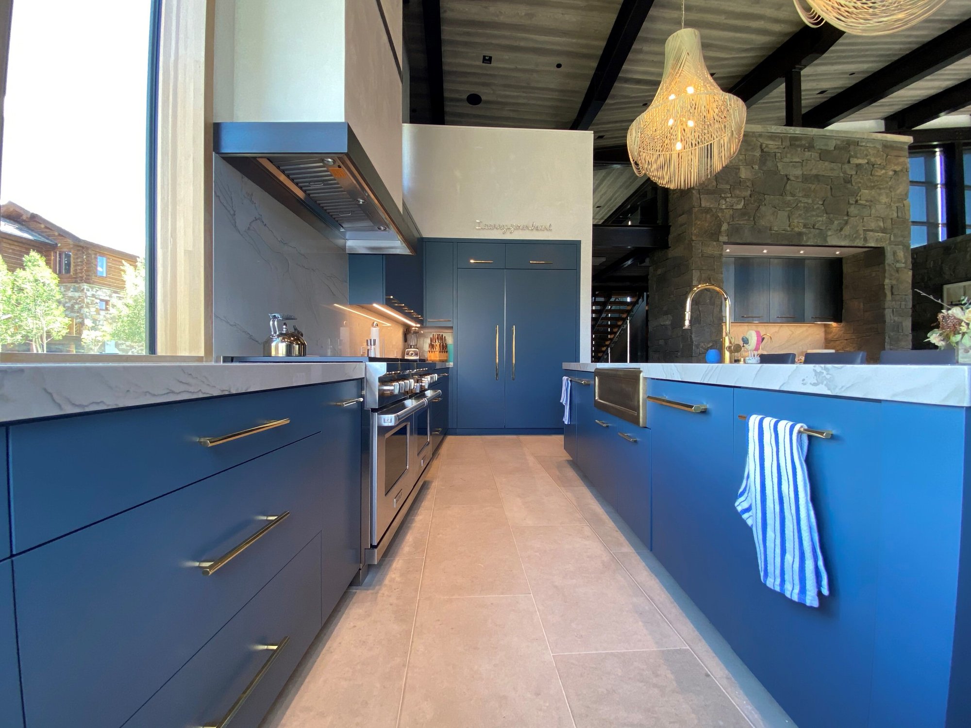 Large trendy u-shaped limestone floor, gray floor and wood ceiling open concept kitchen with a farmhouse sink, flat-panel cabinets, blue cabinets, marble countertops, white backsplash, marble backsplash, paneled appliances, an island and white countertops, by Jensen Design in Telluride, CO