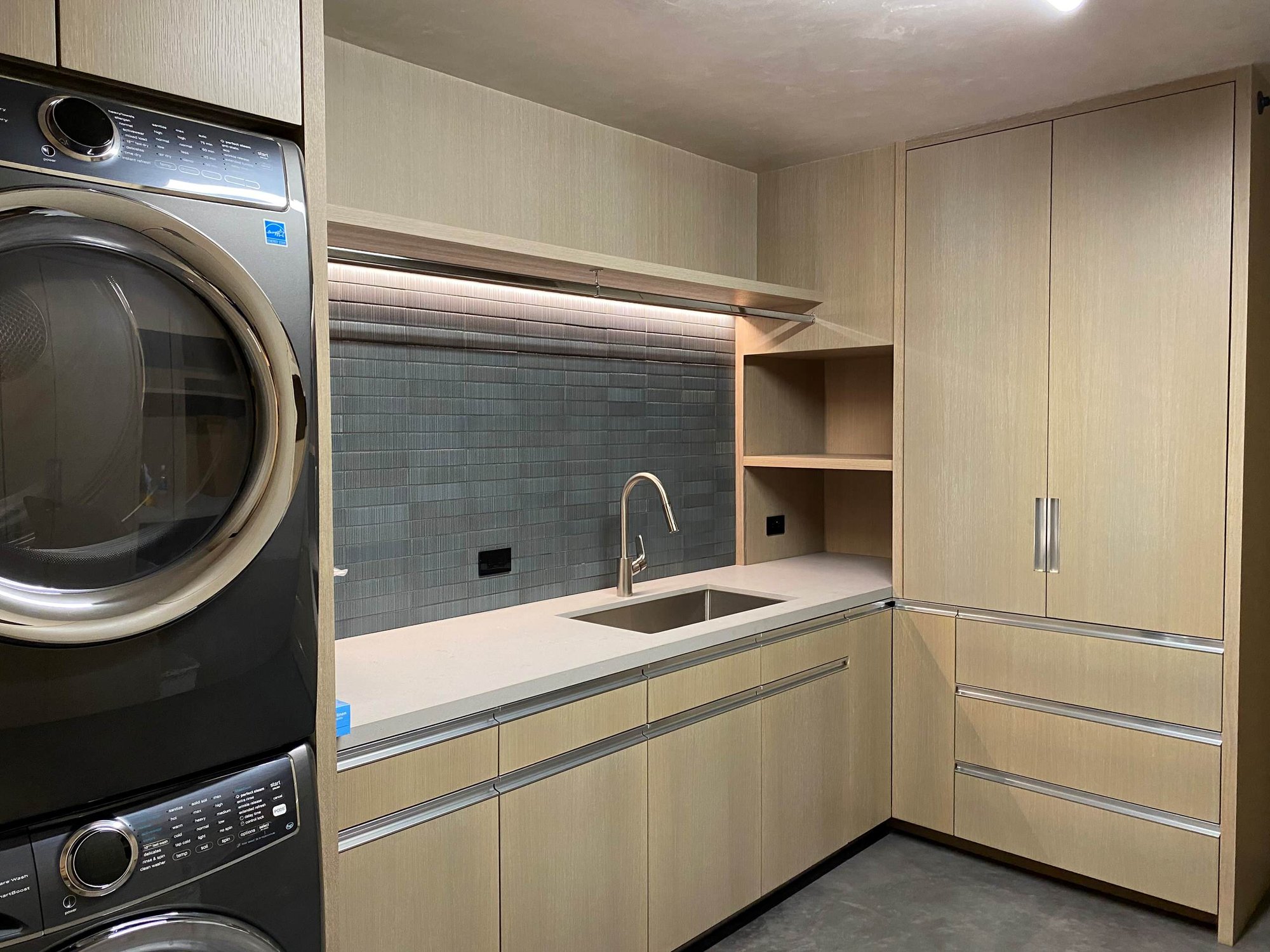 Mid-sized contemporary l-shaped concrete floor, gray floor and wood wall dedicated laundry room with a single-bowl sink, flat-panel cabinets, light wood cabinets, gray backsplash, a stacked washer/dryer and white countertops, by Jensen Design in Telluride, CO