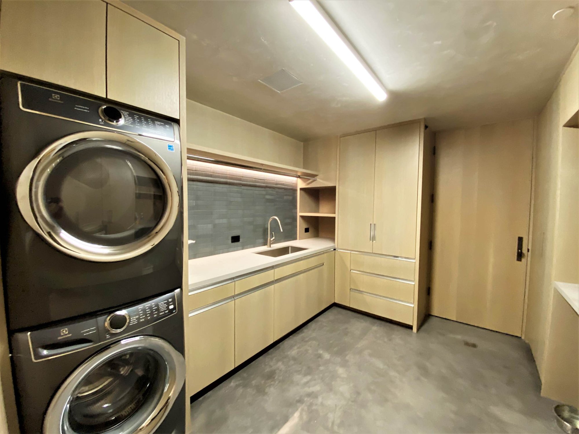 Mid-sized trendy l-shaped concrete floor, gray floor and wood wall dedicated laundry room with a single-bowl sink, flat-panel cabinets, light wood cabinets, gray backsplash, a stacked washer/dryer and white countertops, by Jensen Design in Telluride, CO